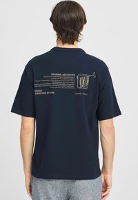 Tengerészkék, rövid ujjú póló, gallérral. A hátsó oldalon fehér szöveg és egy hal konzervben grafikája látható. Az anyag simának és könnyűnek tűnik.