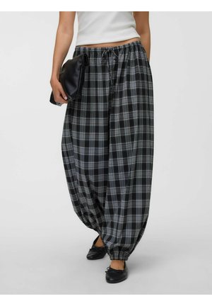Femeie purtând pantaloni evazați în carouri alb-negru cu glezne elastice, un top alb fără mâneci, balerini negri și ținând o poșetă neagră.