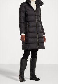 Svart puffjacka med dragkedja framtill och hög krage, med en vadderad design. Parat med svarta leggings och knähöga svarta läderstövlar.