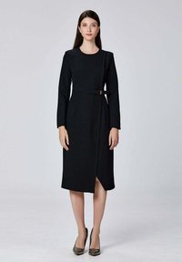 Robe noire à manches longues avec un design habillé, taille ceinturée, silhouette mi-genou et une subtile fente à l'avant. Texture de tissu lisse.