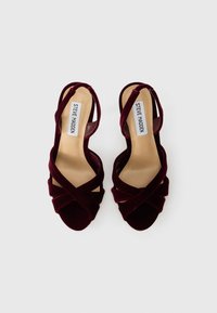 Burgundijske sandale na visokoj peti od baršuna s ukriženim trakama i otvorenim prstima. Unutrašnjost ima bež podlogu s oznakom "STEVE MADDEN".