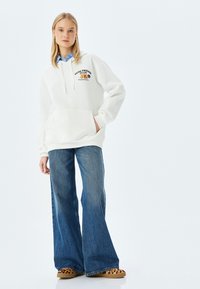 Sudadera blanca con bolsillo frontal y gráfico de "Equipo de Amigos Lindos", combinada con jeans acampanados azules y zapatos tipo slip-on con estampado de leopardo.