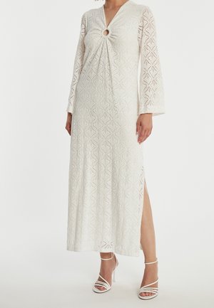 Femme portant une longue robe blanche en dentelle avec des manches cloche, un décolleté en forme de goutte, une fente latérale et des sandales blanches à talons hauts et à brides.
