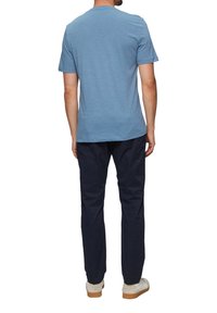Man in een lichtblauwe T-shirt met korte mouwen, donkere marineblauwe broek en witte sneakers, staand met zijn rug naar de camera tegen een witte achtergrond.