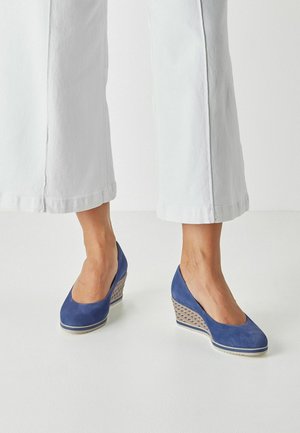 Lábak kék szarvasbőr ék magassarkú cipőben, fehér és barna mintás talppal, fehér felhajtott nadrággal, fehér háttér előtt.