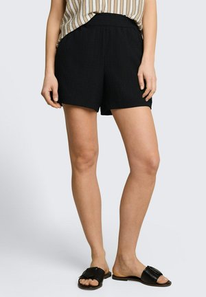 Frau trägt schwarze, strukturierte Shorts, ein beige-weiß gestreiftes Oberteil und schwarze Slip-on-Sandalen und steht vor einem einfarbigen Hintergrund.