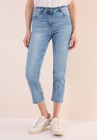 Jeans cropped en denim bleu clair avec un design ajusté, présentant un léger délavage, une taille mi-haute et des détails de couture. Associés à des baskets blanches.