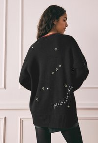 Suéter negro de punto con bordado de estrellas doradas y el texto "falalalala" en la parte posterior. Corte holgado con puños y dobladillo acanalados.