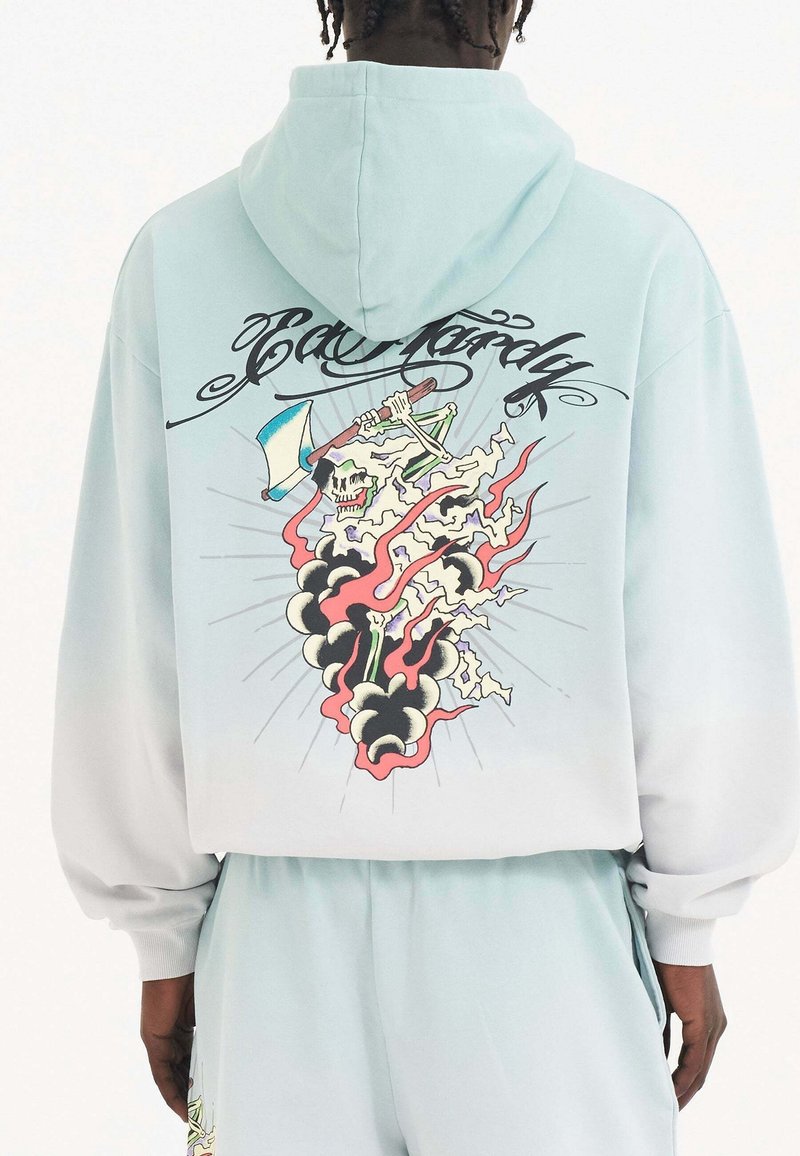 Personne portant un sweat à capuche bleu clair et un pantalon avec un squelette brandissant une hache et des flammes en motif dans le dos, avec un texte stylisé au-dessus.
