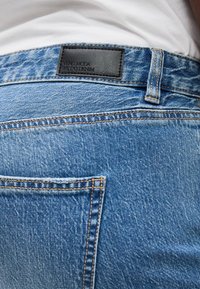 Οπίσθια όψη μπλε τζιν με μαύρη δερμάτινη ετικέτα που γράφει "Vero Moda We Do Denim" πάνω από την τσέπη.