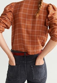 Blouse couleur rouille avec des manches bouffantes et un motif à carreaux blancs, portée avec un jean foncé et une ceinture rouge, vue de dos.