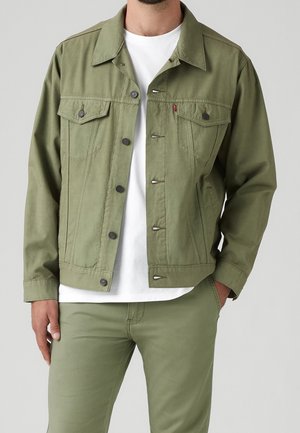 Homme portant une veste boutonnée vert olive sur un t-shirt blanc et un pantalon vert olive assorti, une main dans la poche, fond neutre.