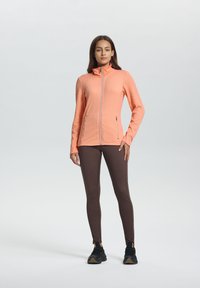 Lichtgewicht, textuurjasje met rits in feloranje, gecombineerd met strakke, bruine leggings en zwarte sportschoenen.