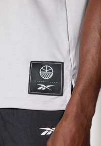 Svetlošedé športové tričko s textúrovaným vzorom, s čiernym štítkom s grafickým znázornením basketbalovej lopty a bielym logom Reebok.
