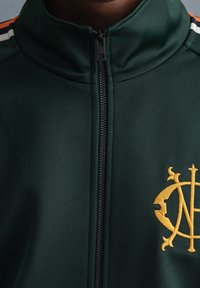 Chaqueta verde con cremallera y alto cuello, con franjas contrastantes en naranja y blanco en los hombros, y un logo bordado en dorado en el pecho.