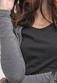 Cardigan gris sur une chemise noire, avec des manches longues et un motif texturé sur les manches. Cheveux ondulés.
