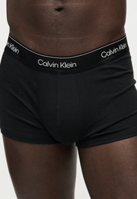 Άνδρας που φοράει μαύρα εσώρουχα Calvin Klein boxer briefs, δείχνοντας τη μέση μέχρι το επάνω μέρος των μηρών σε μονόχρωμο ανοιχτό φόντο.