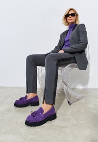Harmaa räätälöity puku, jossa on violetti yläosa, yhdistetty violetteihin mokkanahkaisiin loafer-kenkiin, joissa on frangeja ja paksu musta pohja. Selkeä tuoli on näkyvissä.
