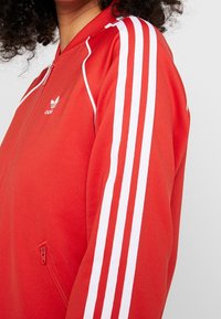 Červená zipová bunda s bílým třemi pruhy na rukávech. Má měkkou látku a malé logo na hrudi. Obsahuje boční zipovou kapsu.