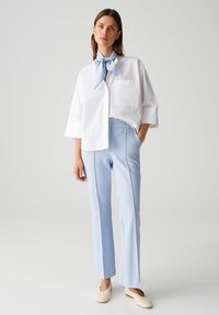 Weißes übergroßes Hemd mit drei Viertelärmeln und Tasche, kombiniert mit hellblauen, taillierten Hosen und cremefarbenen Slippern, akzentuiert mit einem blau gestreiften Schal.