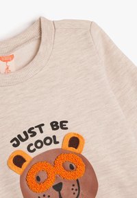 Sudadera beige con un diseño de cara de oso, gafas naranjas y el texto "SOLO SE COOL" en negro. Textura suave, cuello acanalado.