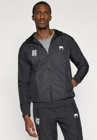 Μαύρο μπουφάν με φερμουάρ και κουκούλα, με υφή σχεδίου στο μπροστινό μέρος και λευκές λεπτομέρειες λογότυπου. Ελαφρύ ύφασμα και ελαστικές μανσέτες.