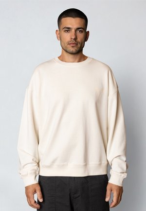 Mężczyzna o krótkich ciemnych włosach, ubrany w luźną, kremową bluzę i czarne spodnie, stojący na tle jednolitego jasnego tła.