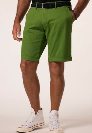 Mann trägt grüne Shorts mit hochgekrempeltem Saum, schwarzen Gürtel, weiße High-Top-Sneaker, steht mit einer Hand in der Tasche vor schlichtem Hintergrund.