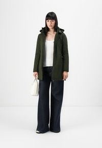 Olijfgroene gewatteerde jas met capuchon, ritsluiting en zijzakken, gecombineerd met hoog uitgesneden donkerblauwe wijde jeans en een witte tas.