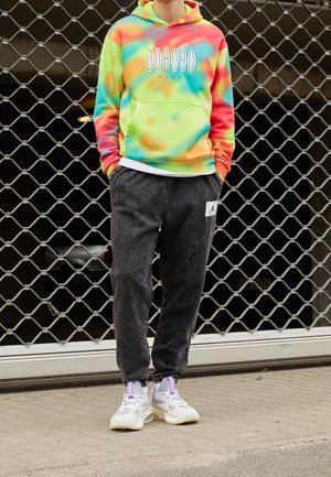 Osoba ubrana w wielokolorową bluzę z kapturem Jordan tie-dye, czarne spodnie dresowe z logo Jordan oraz białe sneakersy z fioletowymi detalami.