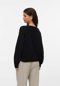 Kvinna med mörkt hår som bär silverfärgade ringörhängen, en svart långärmad tröja och ljusbeiga byxor, stående mot en vit bakgrund.