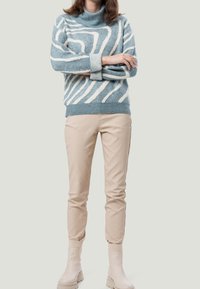 Person, der einen hellblauen und weißen gemusterten Pullover, beige Hosen und beige Knöchelschäfte trägt, steht mit verschränkten Armen vor einem schlichten Hintergrund.