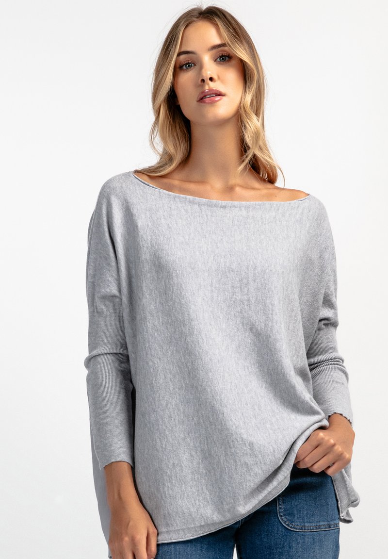 Jasnoszary, luźny sweter z dzianiny z szerokim dekoltem i prążkowanymi rękawami. Miękka faktura; noszony z niebieskimi dżinsami.