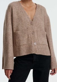 Cárdigan de punto beige con escote en V, tres botones, puños y dobladillo acanalados, y bolsillos de parche frontales. Llevado sobre pantalones de mezclilla negros.