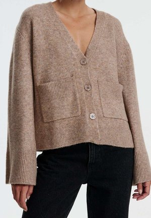 Beige strikket cardigan med V-udskæring, tre knapper, ribbede manchetter og kant, samt forreste påsat lommer. Bæres over sorte denim bukser.