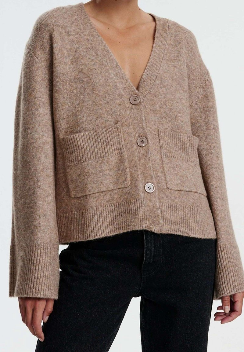 Cárdigan de punto beige con escote en V, tres botones, puños y dobladillo acanalados, y bolsillos de parche frontales. Llevado sobre pantalones de mezclilla negros.