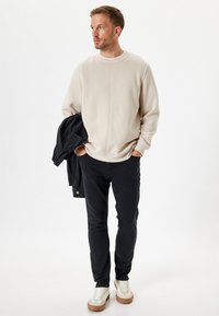 Sudadera beige con cuello redondo y detalles en las costuras, combinada con pantalones negros y una chaqueta oscura, complementada con zapatillas blancas con suelas beige.