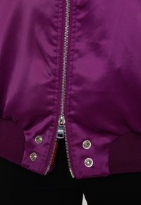 Paarse bomberjack met een glanzende satijnen buitenkant, metallic ritssluiting en ribgebreide manchetten. Oogjes zijn zichtbaar nabij de zoom.