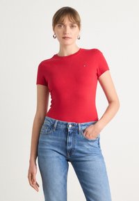 T-shirt rouge à manches courtes au design ajusté, associé à un jean bleu clair. Le t-shirt présente un petit logo sur la poitrine. Texture lisse.