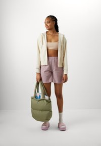 Lys kremfarvet zip-hoodie, beige crop top, lyserøde teksturerede shorts og en overdimensioneret olivengrønn taske. Lyserøde platformsandaler med hvide sokker.