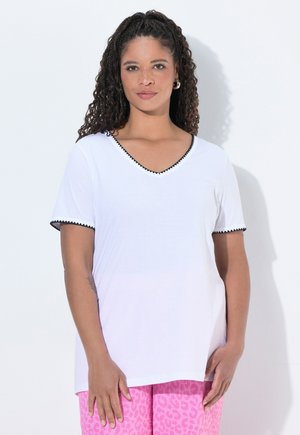 Mujer con cabello rizado que lleva una camiseta blanca de cuello en V con ribete negro y pantalones rosas con estampado de leopardo, de pie frente a un fondo blanco.