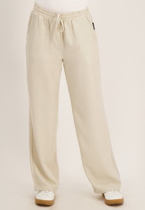 Beige sweatpants met wijde pijpen, een elastische tailleband en trekkoord, gedragen met witte sneakers met rubberen zolen.