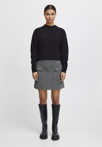Pull en maille noire, jupe en tweed gris avec poches plaquées, et bottes noires en cuir à hauteur de genou. Silhouette simple et structurée ; tissu texturé.