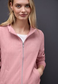 Ung kvinde iført gullige hoop-øreringe og en rosa lynlåset ribbet jakke over en hvid skjorte, der står mod en mørkegrå baggrund.