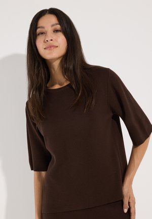 Frau mit langen dunklen Haaren, die ein lockeres braunes Kurzarmshirt trägt und vor einem einfarbigen hellen Hintergrund steht.