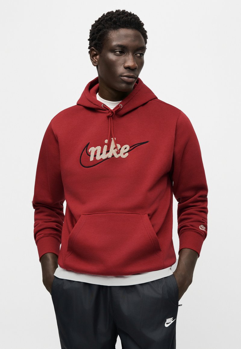 Felpa rossa con cappuccio e tasca a marsupio, con un logo "Nike" ricamato beige. Tessuto morbido e polsini a coste. Indossata sopra una camicia bianca.