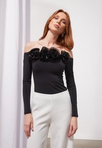 Juodas off-the-shoulder bodysuit'as su gėlių nėriniais apykaklėje, ilgomis rankovėmis, derintas su baltais plačiais kelnais.