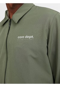Veste verte avec un col et une fermeture à glissière à l'avant. Présente le logo "core dept." et les coordonnées géographiques imprimées sur la zone de la poitrine gauche. Texture lisse.
