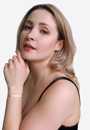 Bracciale d'oro con un ciondolo rettangolare, texture liscia, indossato su un polso, su uno sfondo neutro, abbinato a una maglietta nera.