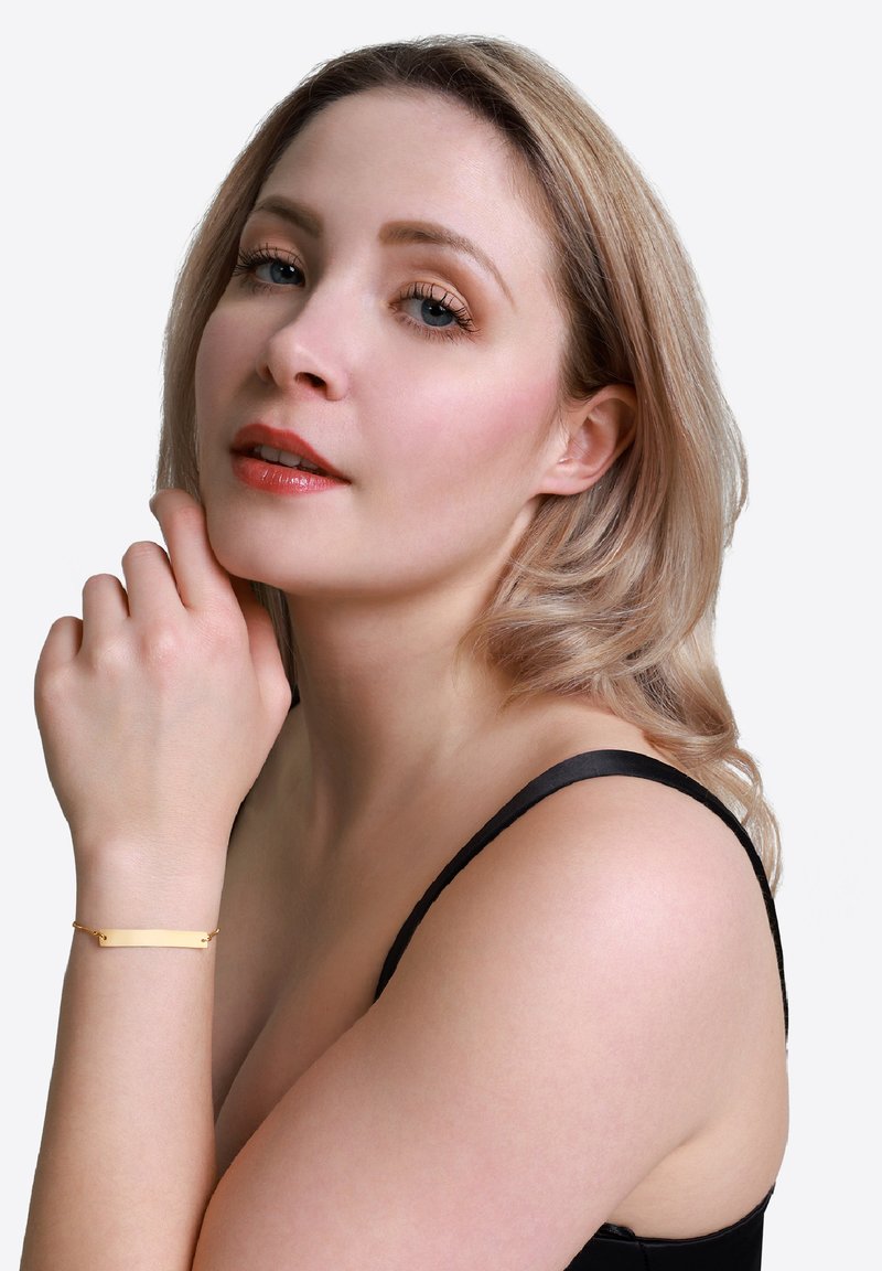 Bracciale d'oro con un ciondolo rettangolare, texture liscia, indossato su un polso, su uno sfondo neutro, abbinato a una maglietta nera.